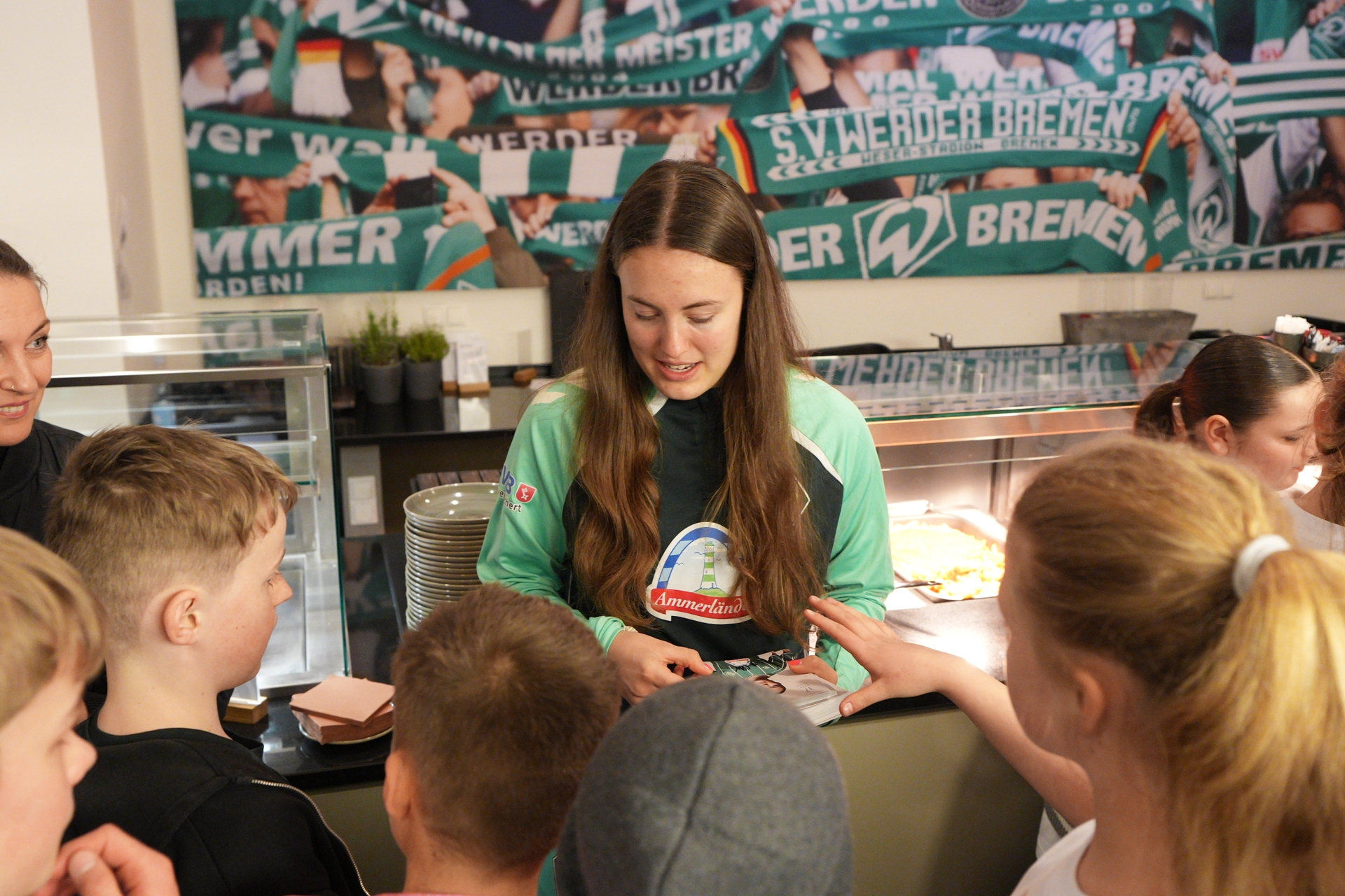 Eine junge Frau in einem grünen Trikot gibt Kindern Autogramme, im Hintergrund sind Sportbanner zu sehen.