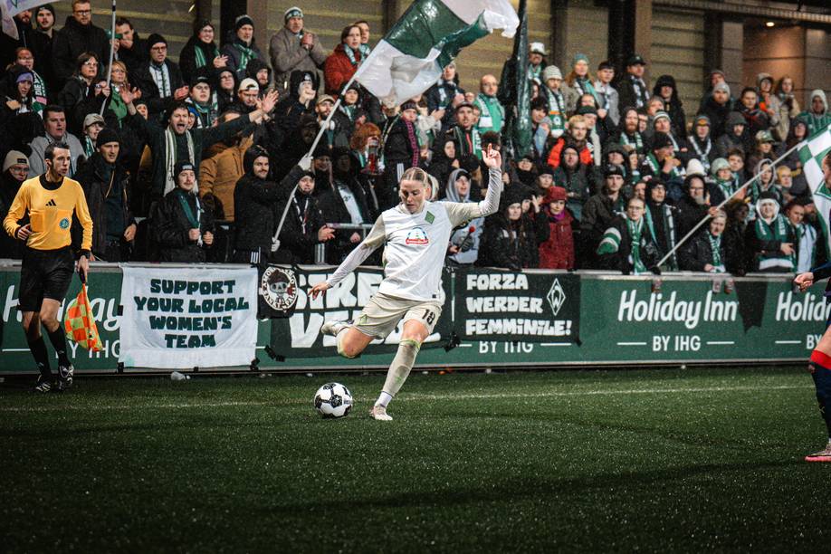 Lina Hausicke in weißem Trikot schießt den Ball auf einem Spielfeld, im Hintergrund jubelt die Menge und es sind Fanbanner zu sehen.