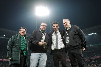 Vier Männer stehen auf einem Fußballfeld unter hellen Stadionlichtern und tragen Jacken und Schals. Einer hält eine dreieckige Auszeichnung in der Hand.