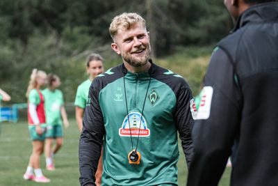 Ein lächelnder Mann mit Bart, der einen grünen Sportpullover mit Logo trägt und eine Trillerpfeife um den Hals hat; er steht im Freien, im Hintergrund sind verschwommene Menschen zu sehen.