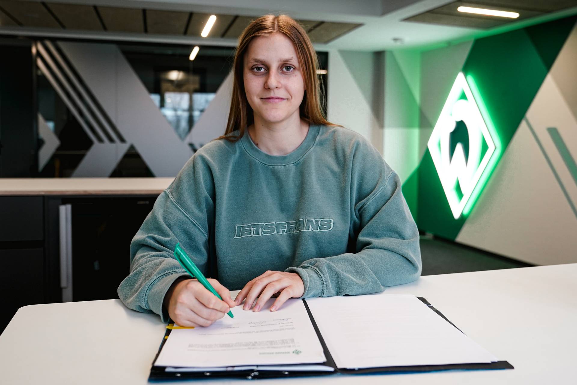 Sarah Gutmann sitzt an einem weißen Tisch. Vor ihr liegt eine Vertragsmappe, sie hat einen Stift in der Hand. Im Hintergrund leuchtet eine Werder-Raute.