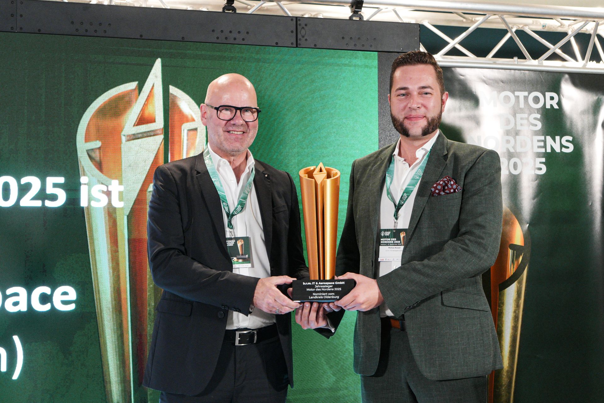 Zwei Männer, einer mit Glatze und einer mit dunklen Haaren, stehen im Anzug nebeneinander und halten gemeinsam einen Pokal fest. Sie lächeln in die Kamera. Im Hintergrund ist eine Bühne zu erkennen.