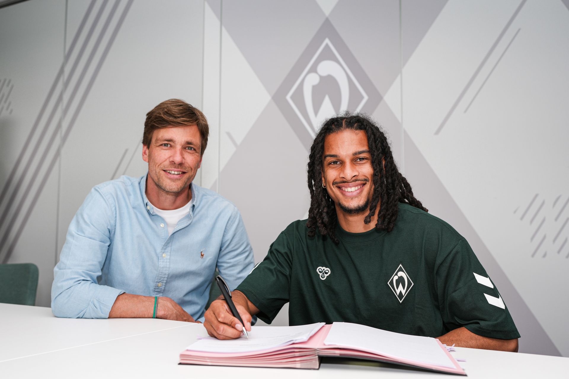 Clemens Fritz und Isaac Schmidt sitzen am Tisch. Schmidt unterschreibt seinen Vertrag. Im Hintergrund ist eine Werder-Raute an der Wand.