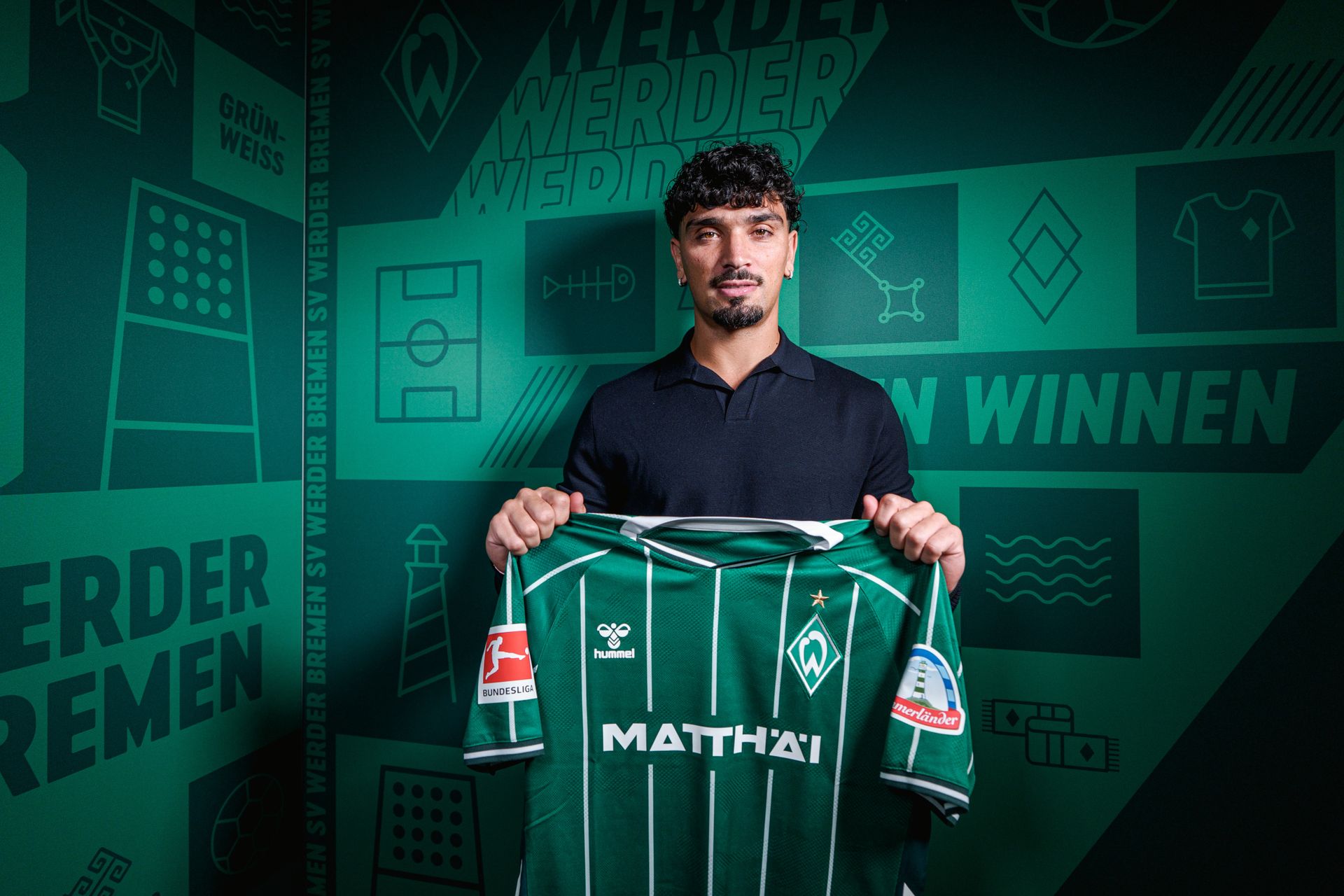 Cameron Puertas hält ein Werder-Trikot hoch. Im Hintergrund ist ein Werder-Banner.