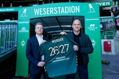 Stephan Ehmke (HAWE) und Klaus Filbry halten das dritte Trikot der Saison 2025/26 in den Händen. Auf dem Rücken ist ein Flock aufgedruckt: "WERDER 26/27 Hanseatische Weinhandelsgesellschaft". Sie stehen vor dem Eingang des Spielertunnels im Weserstadion.
