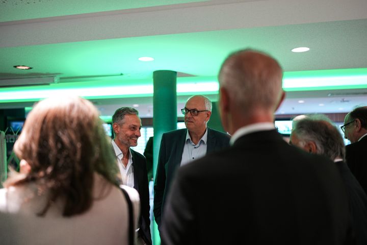 Das Foto ist über den Rücken von zwei Personen aufgenommen und fokussiert zwei Männer mittleren Alters, die in einem Kreis stehen und sich unterhalten.