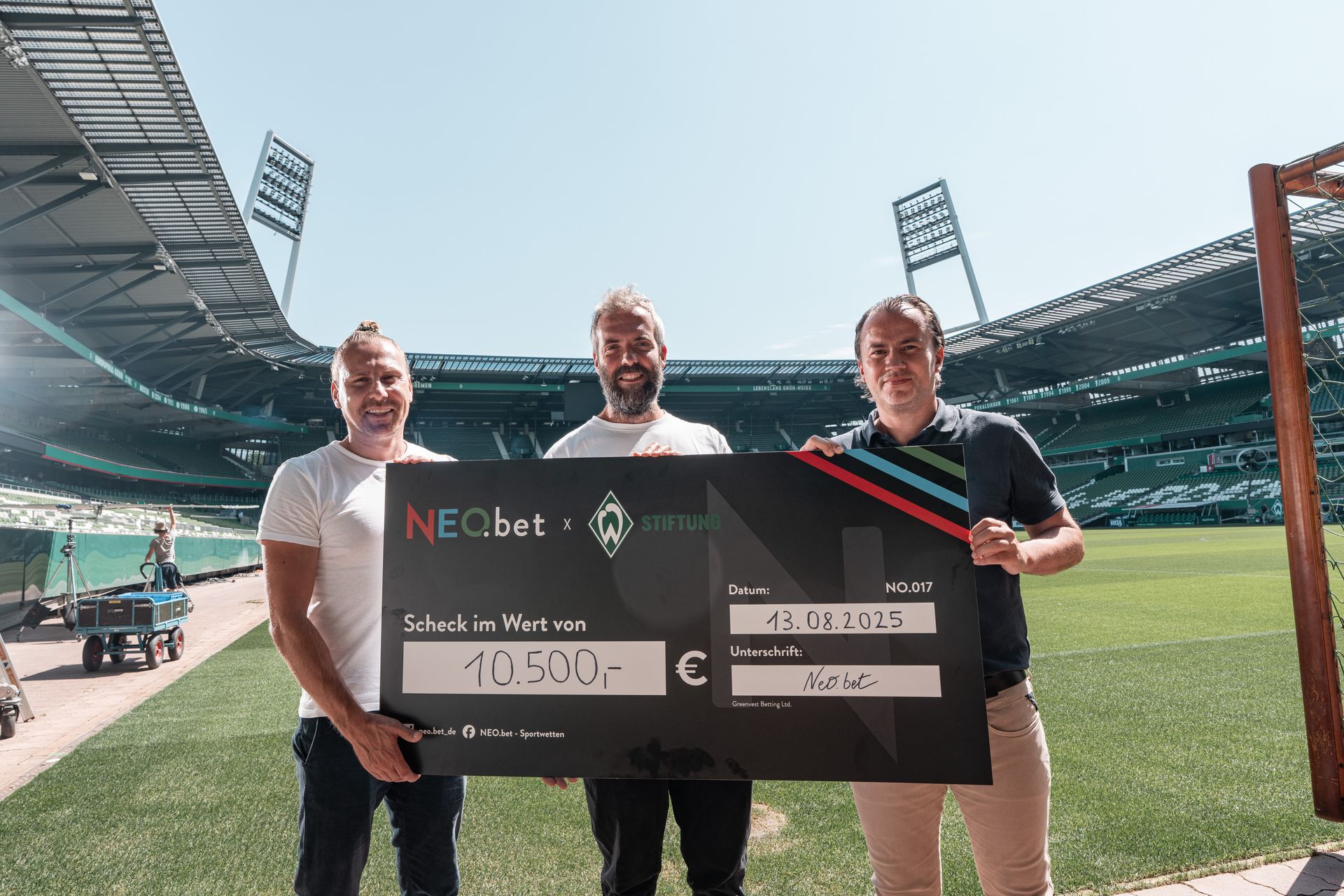 Drei Männer stehen auf einem Fußballfeld und halten einen großen Scheck über 10.500 Euro in den Händen. Im Hintergrund ist das Stadion zu sehen.