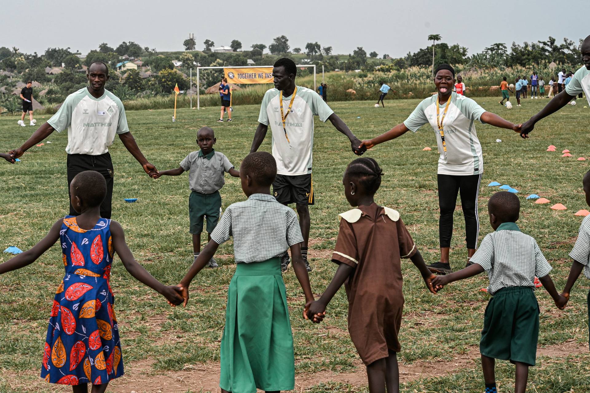 Kinder und Erwachsene stehen Hand in Hand im Kreis auf einer Wiese und nehmen an einer Gruppenaktivität oder einem Spiel im Freien teil.