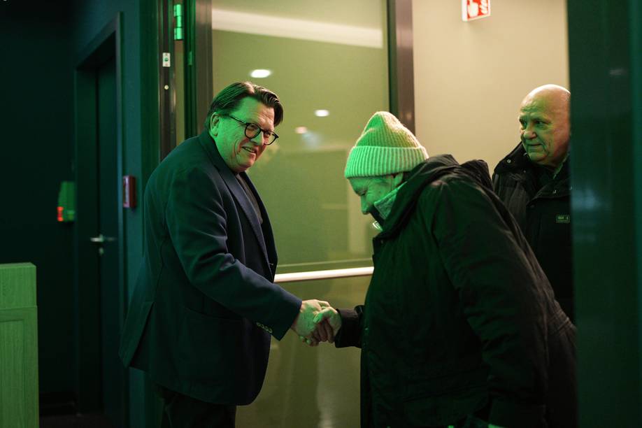 Ein Mann in einer blauen Jacke gibt einem anderen Mann mit einer Mütze die Hand, während eine dritte Person in einem grün beleuchteten Raum zusieht.