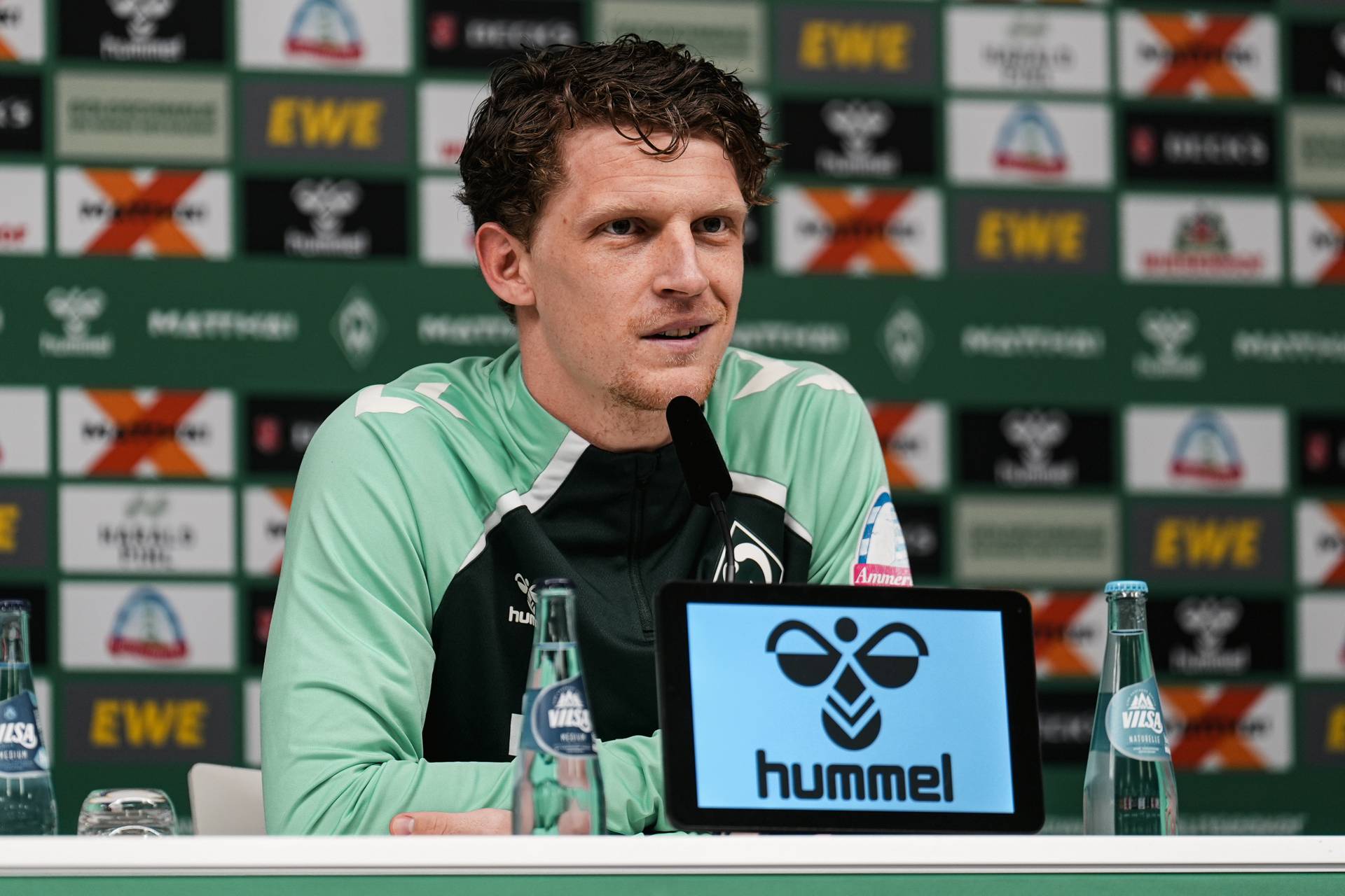 Jens Stage sitzt im grünen Shirt vor einer Sponsorenwand und spricht in ein Mikrofon. Vor ihm steht ein Tablet, worauf in schwarz das hummel-Logo auf weißem Grund zu sehen ist.