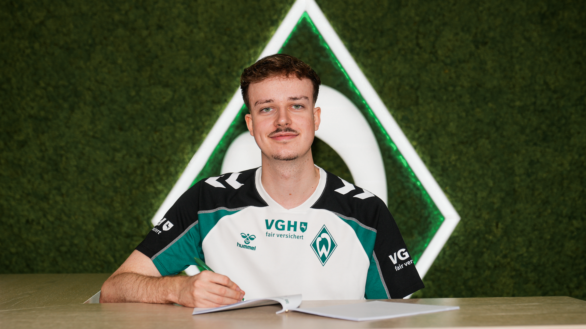 Max Julius "Diviners" Gröne in einem Sportshirt sitzt lächelnd an einem Tisch vor einem grünen Hintergrund mit einem Werder Bremen Logo.