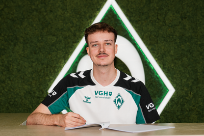Max Julius "Diviners" Gröne in einem Sportshirt sitzt lächelnd an einem Tisch vor einem grünen Hintergrund mit einem Werder Bremen Logo.