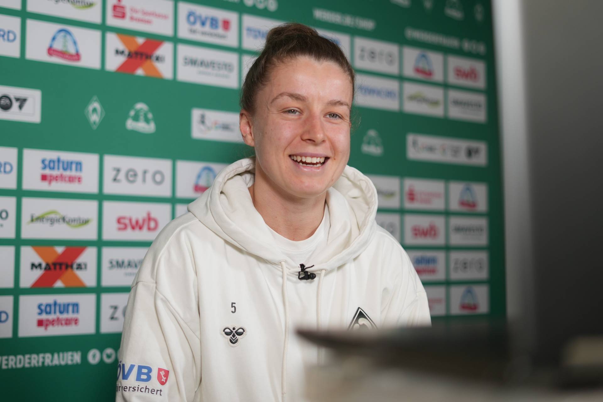 Michelle Ulbrich lacht. Sie trägt einen hellen Pullover und sitzt vor einer grün-weißen Sponsorenwand.