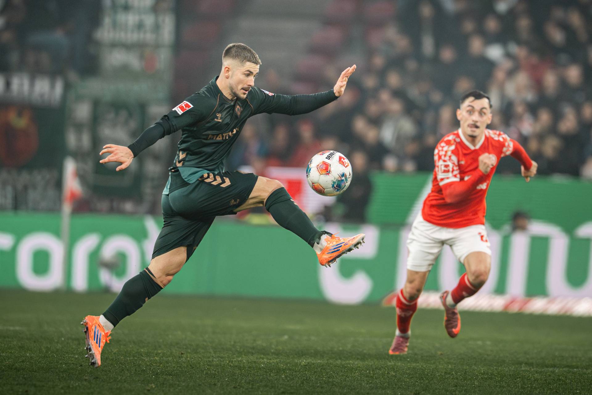 Romano Schmid im dunkelgrünen Trikot springt mit gestrecktem Bein zum Ball, ein Mainzer Spieler in rot läuft im Hintergrund auf ihn zu.