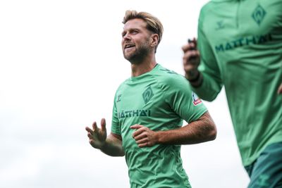 Ein Mann in einem grünen Sporttrikot, der im Freien joggt; rechts ist eine weitere Person teilweise zu sehen.