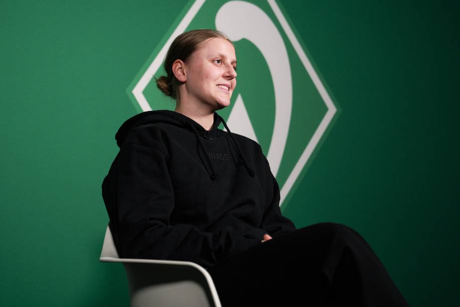 Sarah Gutmann sitzt auf einem weißen Stuhl vor einer grünen Wand. Auf der Wand ist die Werder-Raute zu sehen.