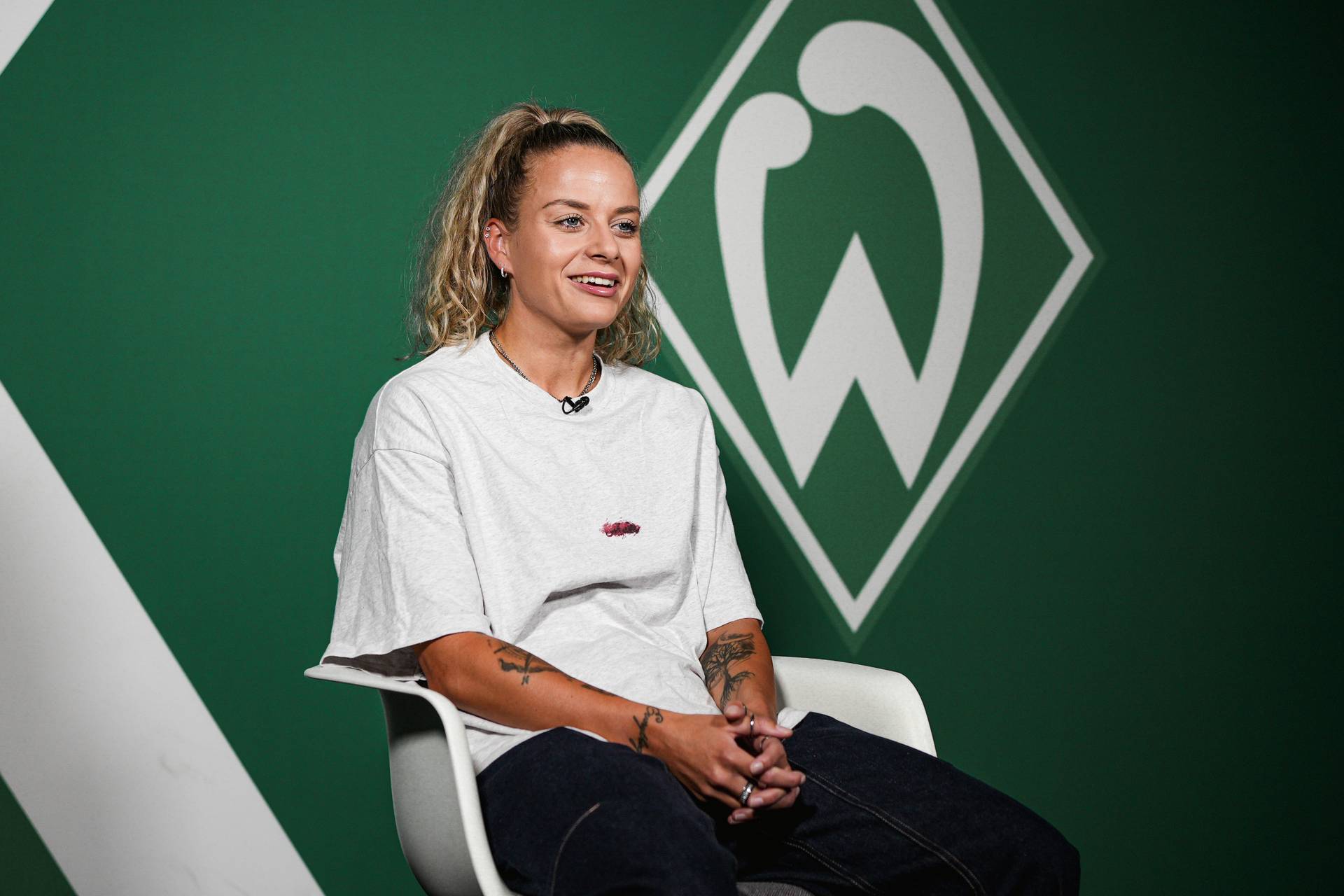 Eine Person mit Pferdeschwanz, die ein weißes T-Shirt und Jeans trägt, sitzt vor einem grünen Hintergrund mit einem stilisierten Logo und lächelt.