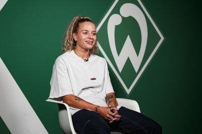 Eine Person mit Pferdeschwanz, die ein weißes T-Shirt und Jeans trägt, sitzt vor einem grünen Hintergrund mit einem stilisierten Logo und lächelt.