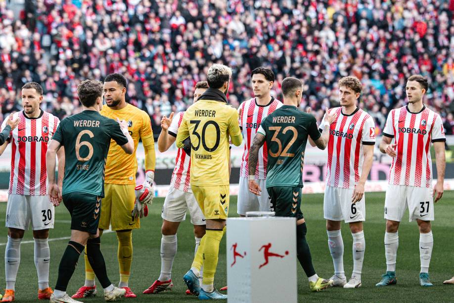 Fußballspieler in rot-weiß gestreiften Trikots geben sich vor einem Spiel auf dem Spielfeld die Hand mit Spielern in grünen Trikots, im Hintergrund ist eine Menschenmenge zu sehen.