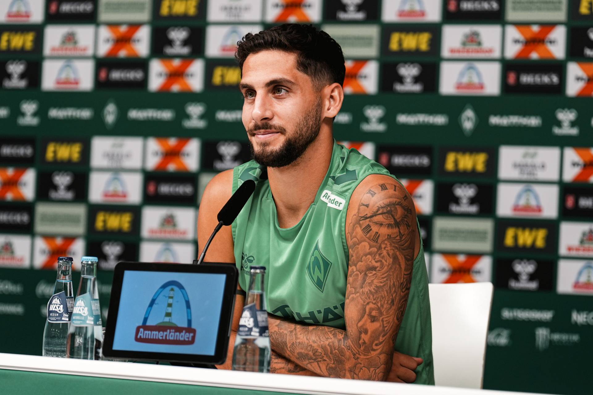 Ein Sportler in einem grünen Shirt sitzt mit einem Mikrofon am Tisch einer Pressekonferenz; sein tätowierter Arm ist zu sehen. Im Hintergrund sind mehrere Sponsorenlogos zu sehen.