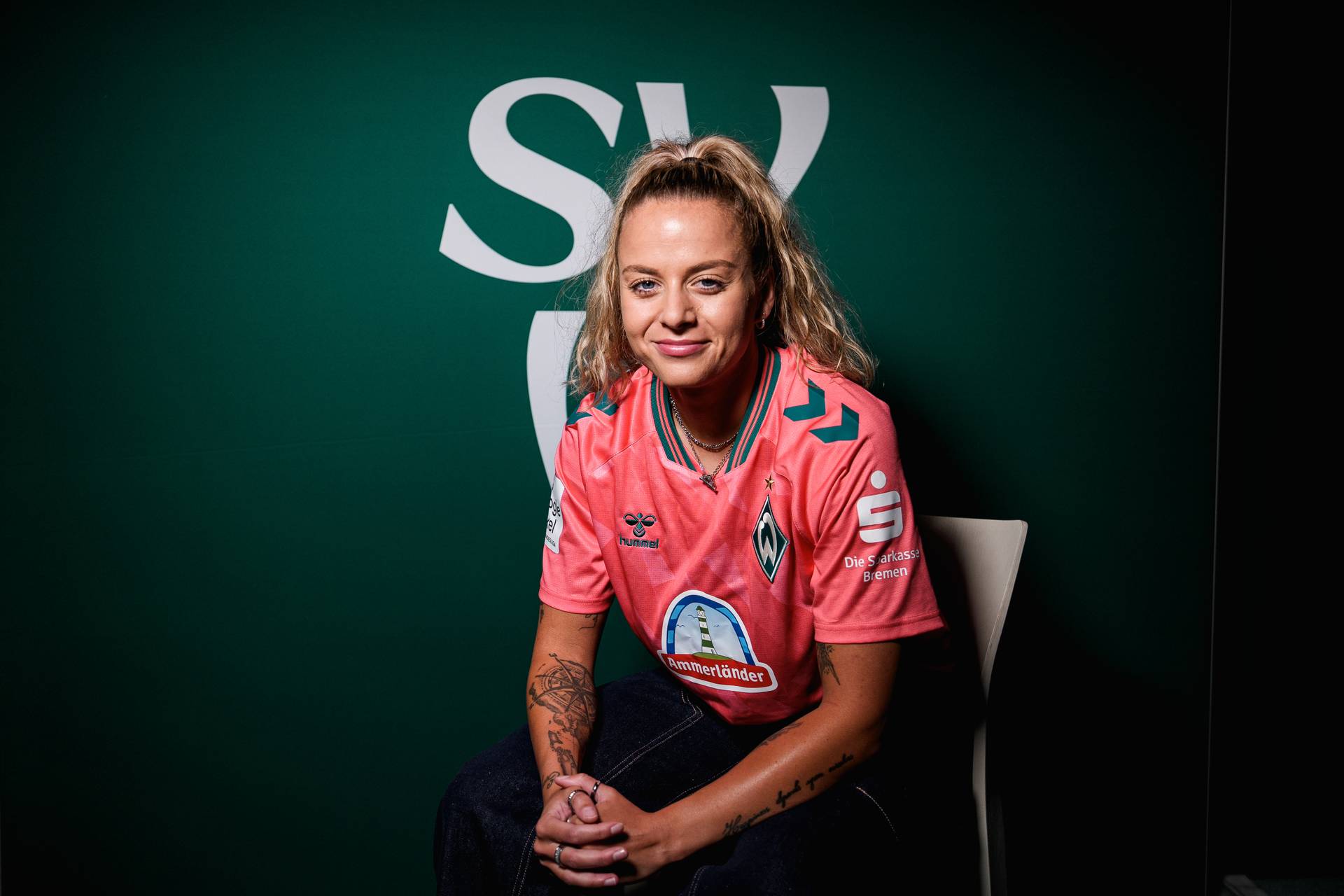 Eine Frau mit Tattoos sitzt in einem rosa Fußballtrikot mit einem Sponsorenlogo vor einem grünen Hintergrund, auf dem in großen weißen Buchstaben „SV“ zu lesen ist.