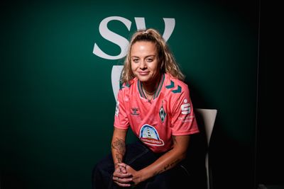 Eine Frau mit Tattoos sitzt in einem rosa Fußballtrikot mit einem Sponsorenlogo vor einem grünen Hintergrund, auf dem in großen weißen Buchstaben „SV“ zu lesen ist.