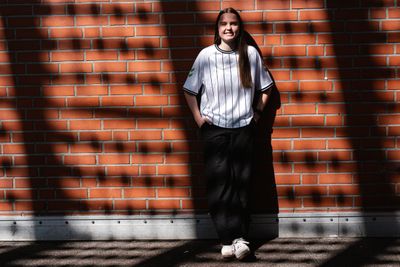 Eine Person in einem weißen Nadelstreifenhemd steht lächelnd vor einer sonnenbeschienenen Backsteinmauer, auf der Schatten ein Muster zeichnen.