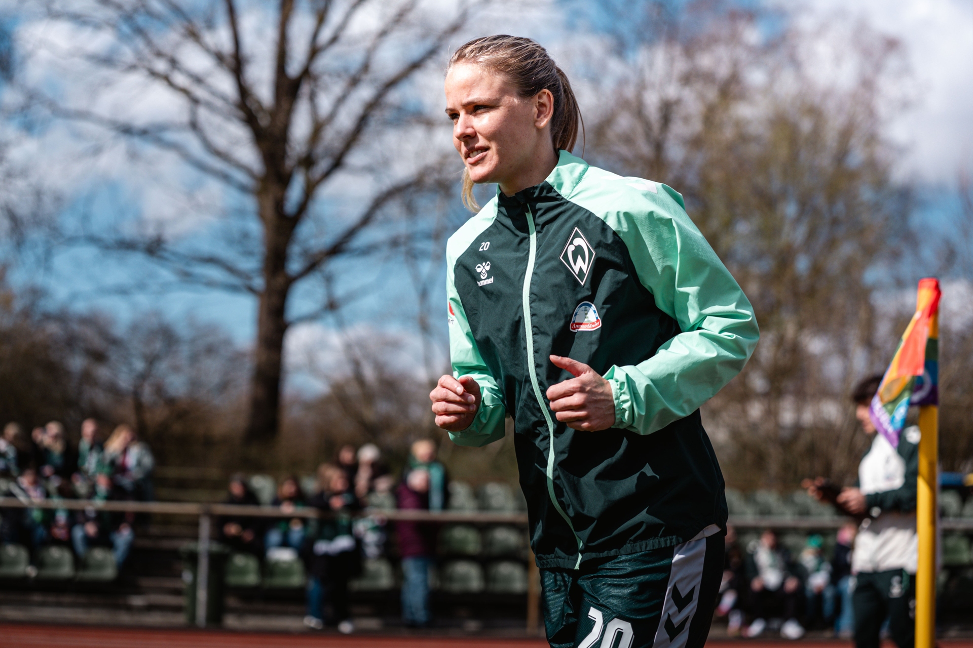 Ein Fußballspieler in einer grün-blaugrünen Jacke joggt auf einem Spielfeld, im Hintergrund sind Zuschauer zu sehen und eine Regenbogenfahne ist zu erkennen.