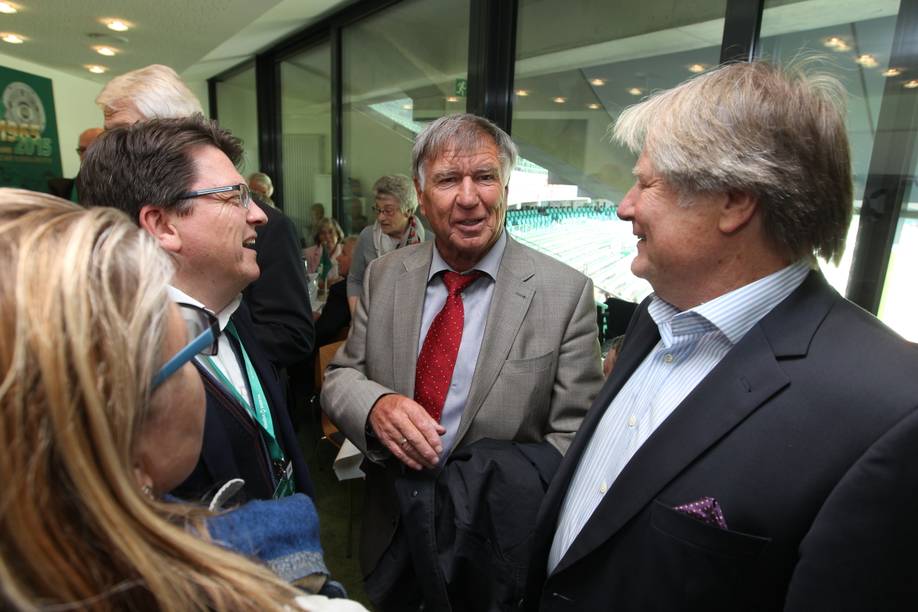 Sepp Piontek in der Mitte mehrerer Menschen. Er schaut in die Kamera. Im Hintergrund ist ein Fußballstadion mit grünen Sitzen zu sehen.