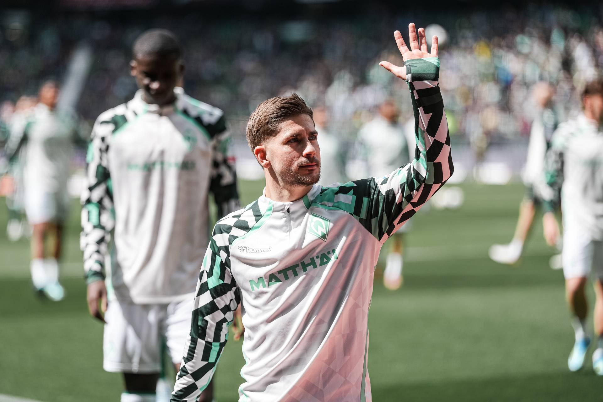Ein Fußballspieler in einem grün-weißen Trikot mit geometrischem Muster winkt den Fans zu, während er über das Spielfeld läuft; im Hintergrund sind seine Mannschaftskameraden zu sehen.