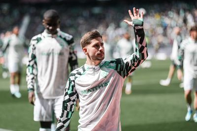 Ein Fußballspieler in einem grün-weißen Trikot mit geometrischem Muster winkt den Fans zu, während er über das Spielfeld läuft; im Hintergrund sind seine Mannschaftskameraden zu sehen.