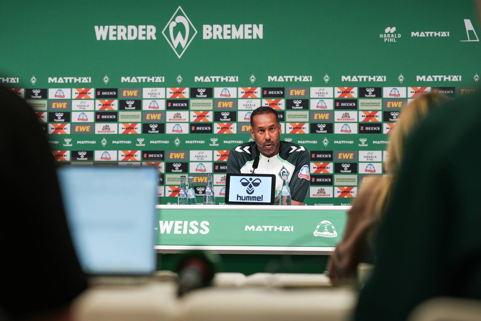 Ein Mann an einem Pressekonferenztisch, im Hintergrund sind Sponsorenlogos zu sehen, außerdem ein Mikrofon und Wasserflaschen.