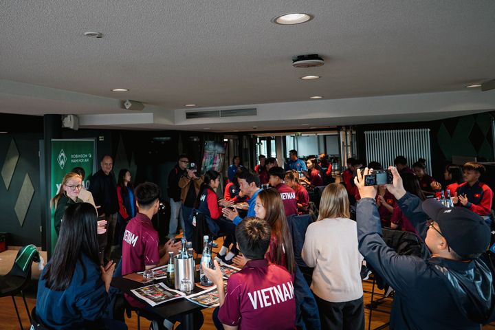 Eine Gruppe von Menschen in einem Raum, von denen einige „Vietnam“-T-Shirts tragen, unterhalten sich und machen Fotos. Es sind Tische mit Flaschen und Papieren zu sehen.