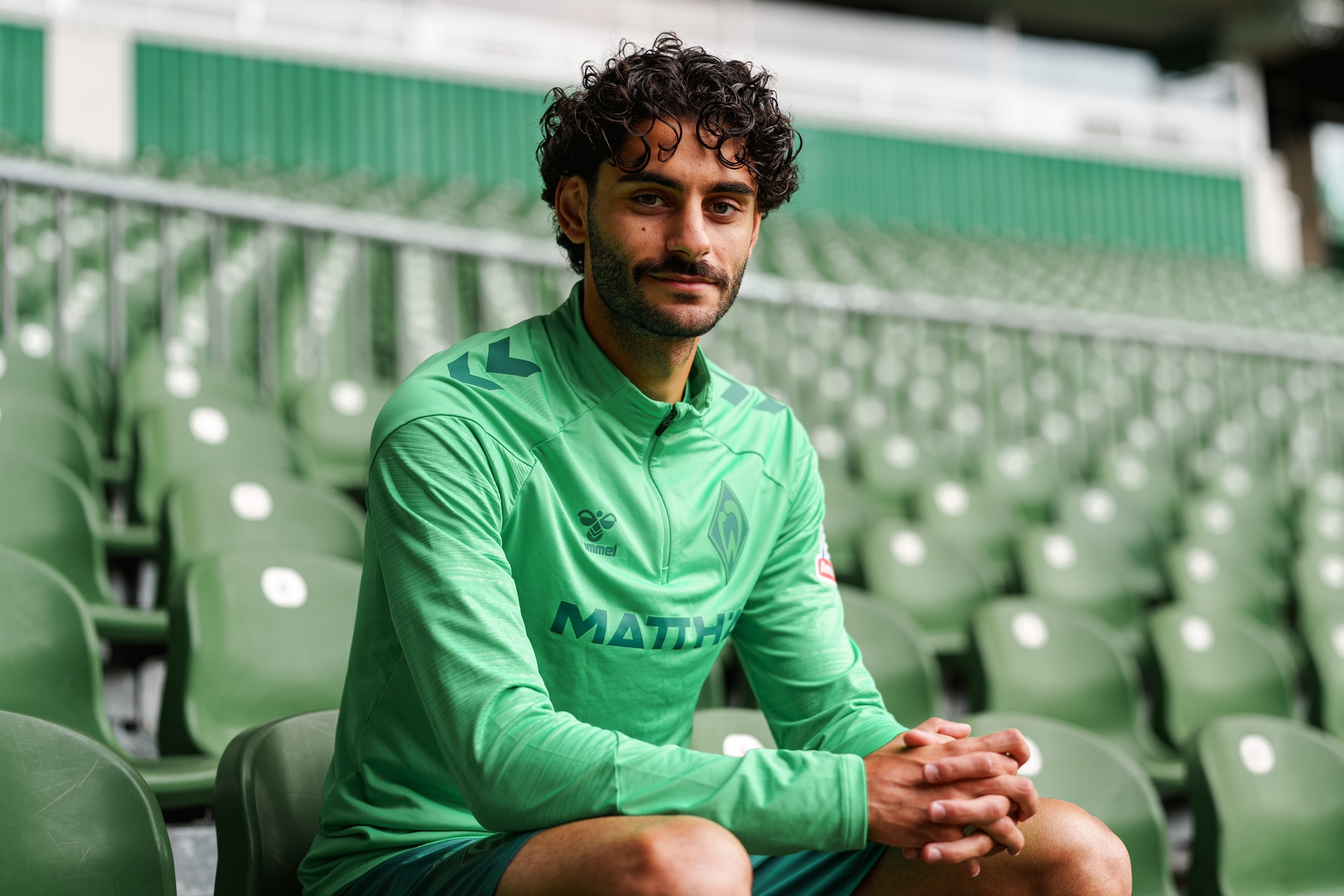 Ein Mann in einem grünen Sporttrikot sitzt auf einem Stadionplatz, umgeben von leeren grünen Sitzen, und blickt in die Kamera.