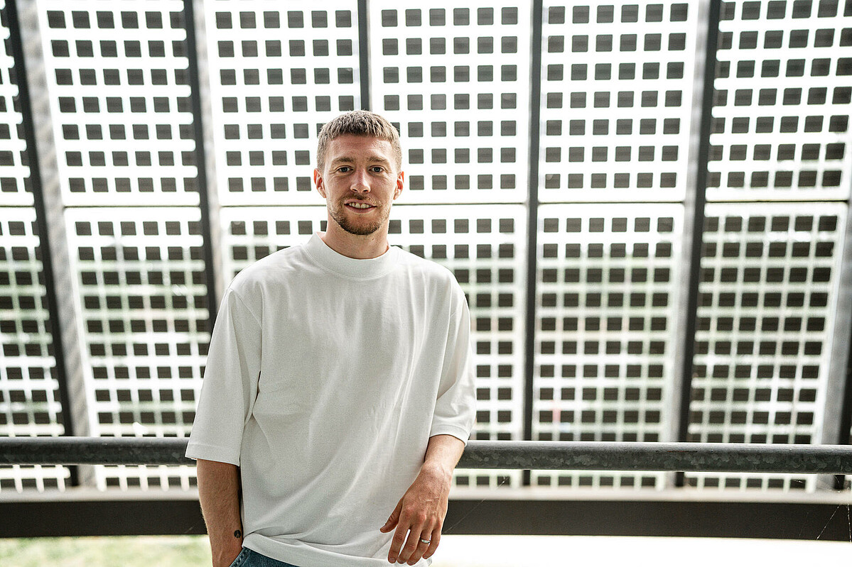null Mitchell Weiser steht vor der Solaranlage am wohninvest WESERSTADION. Er lehnt an ein Geländer. er trägt ein weißes Tshirt und eine blaue Jeansshorts.