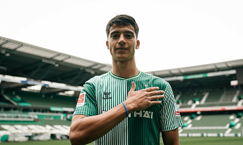 Julian Malatini steht im wohninvest WESERSTADION. Er schaut in die Kamera. Die rechte Hand hat er auf die Werder-Raute gelegt.