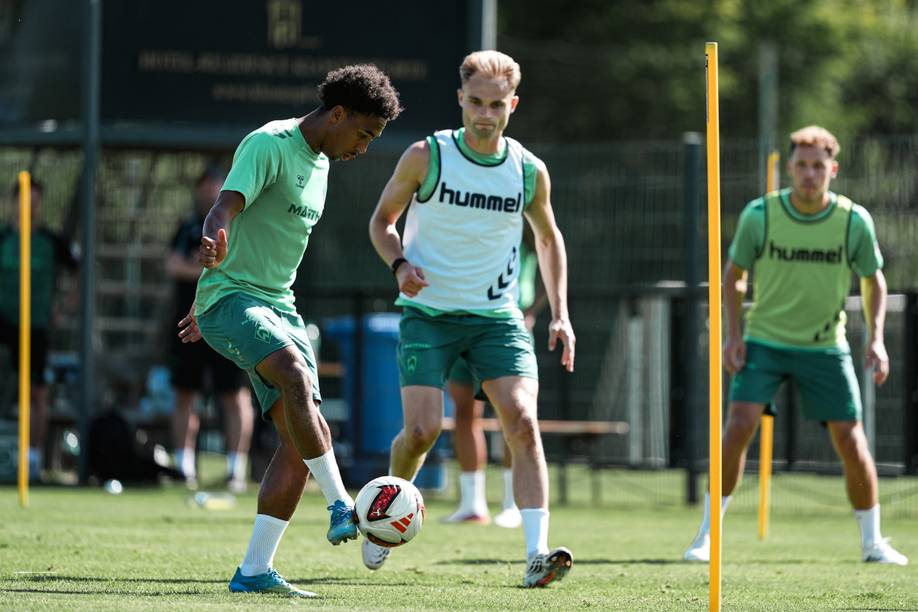 Fußballspieler in grünen Trikots trainieren auf einem sonnigen Platz; einer spielt mit dem Ball, während die anderen zuschauen und warten, im Hintergrund sind Trainingsstangen zu sehen.