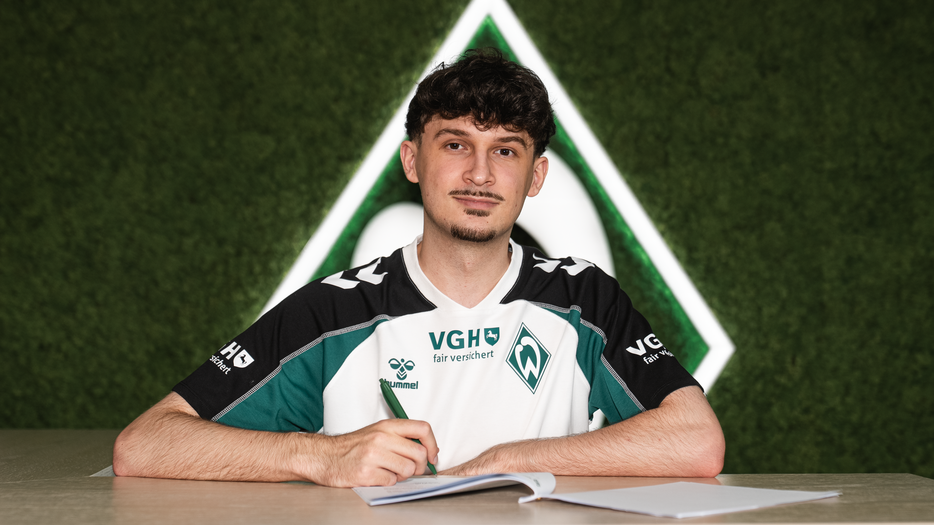 Jannik "Kynuru" Schüßler in einem Sportshirt sitzt lächelnd an einem Tisch vor einem grünen Hintergrund mit einem Werder Bremen Logo.