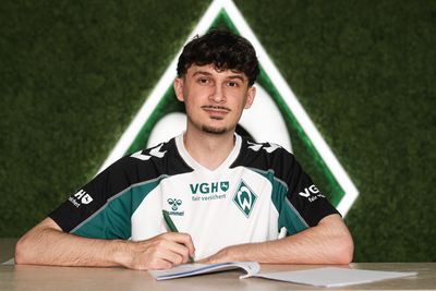Jannik "Kynuru" Schüßler in einem Sportshirt sitzt lächelnd an einem Tisch vor einem grünen Hintergrund mit einem Werder Bremen Logo.