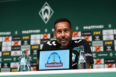 Daniel Thioune sitzt bei der Pressekonferenz vor einem Mikrofon und lächelt.