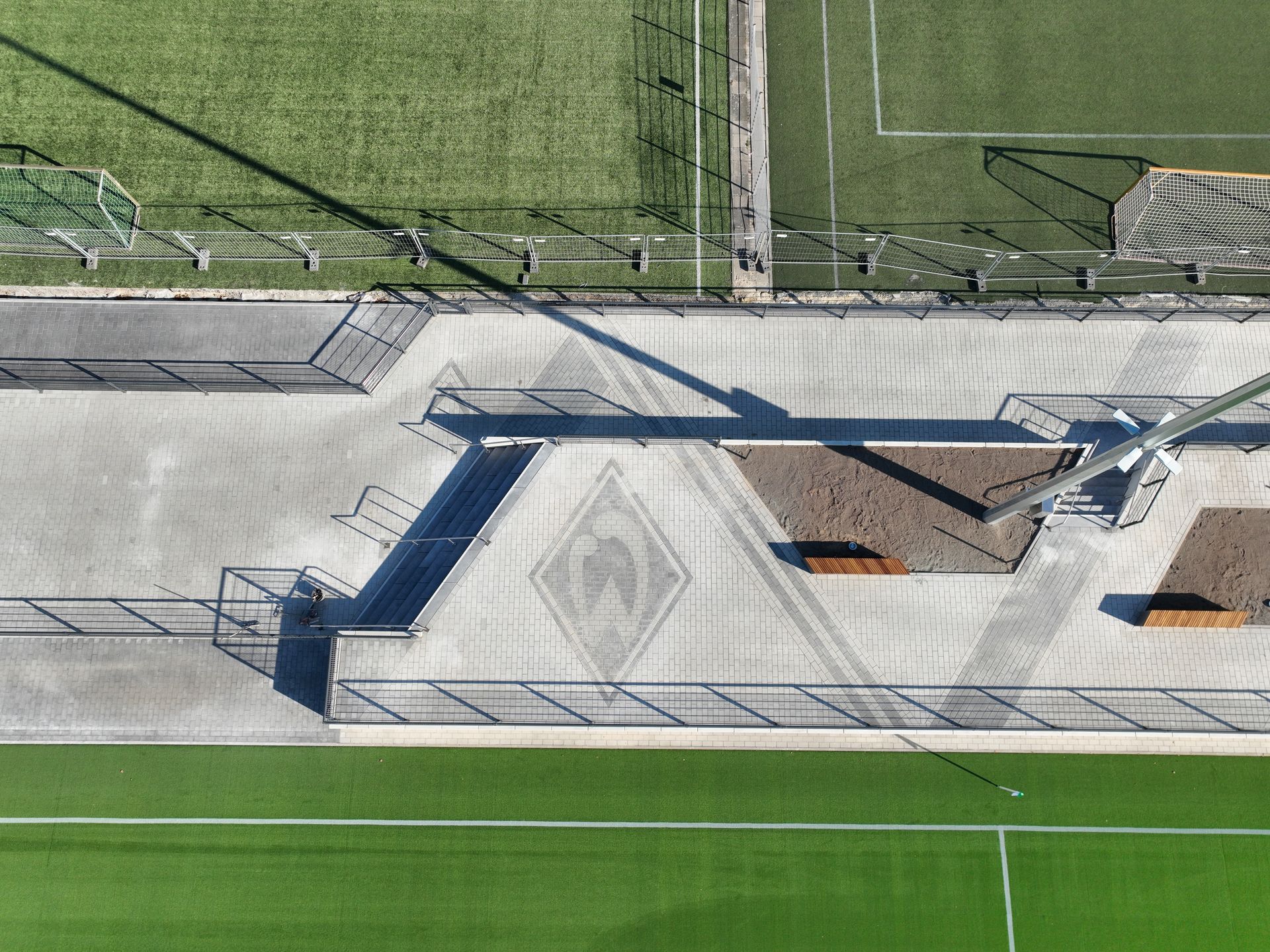 Luftaufnahme eines Sportplatzes mit grünem Kunstrasen, angrenzenden Betonwegen und eingezäunten Bereichen, die lange Schatten werfen.