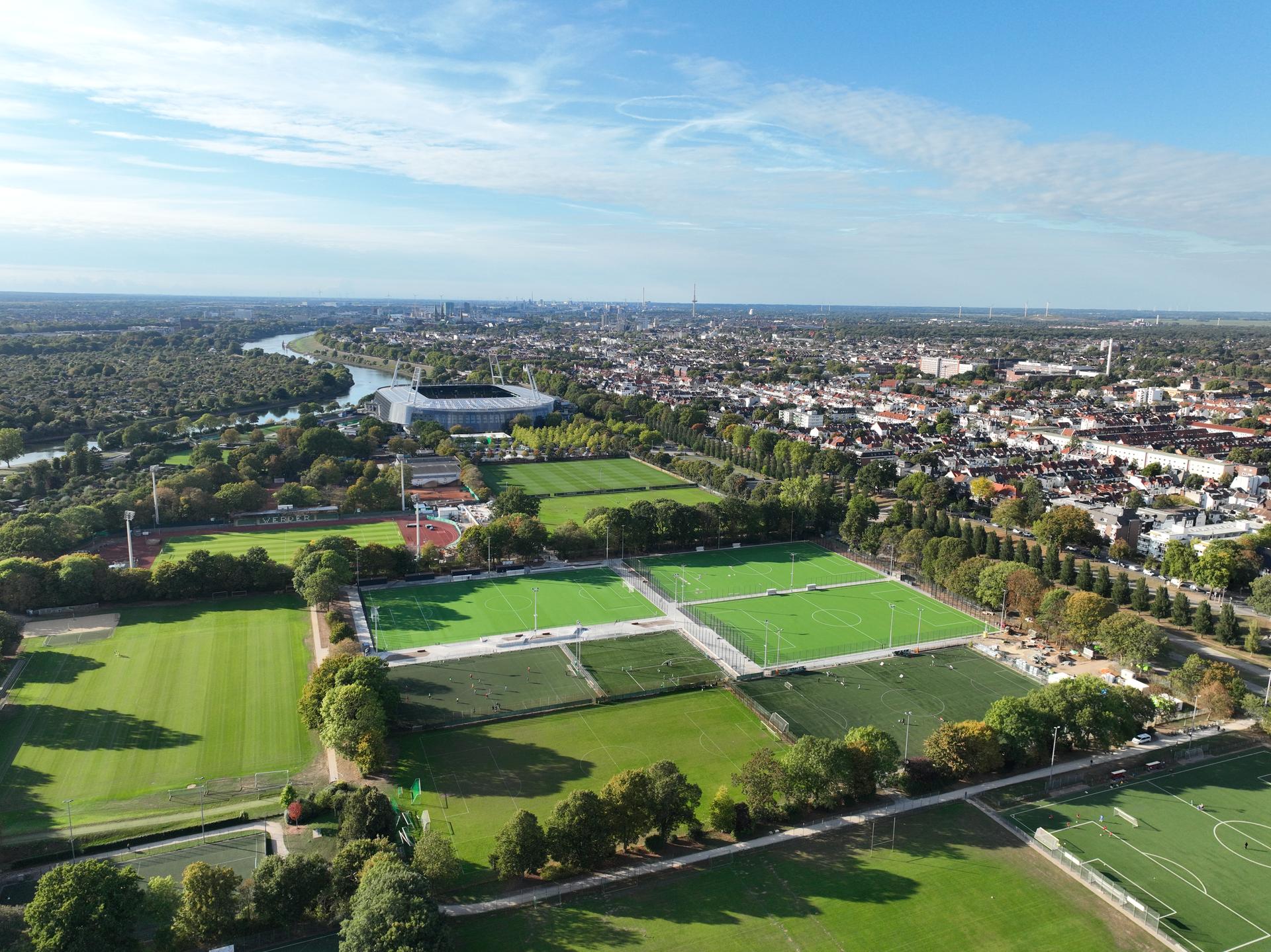 Luftaufnahme einer Stadtlandschaft mit Sportplätzen, einem Fluss und einem von Bäumen umgebenen Stadion unter strahlend blauem Himmel.