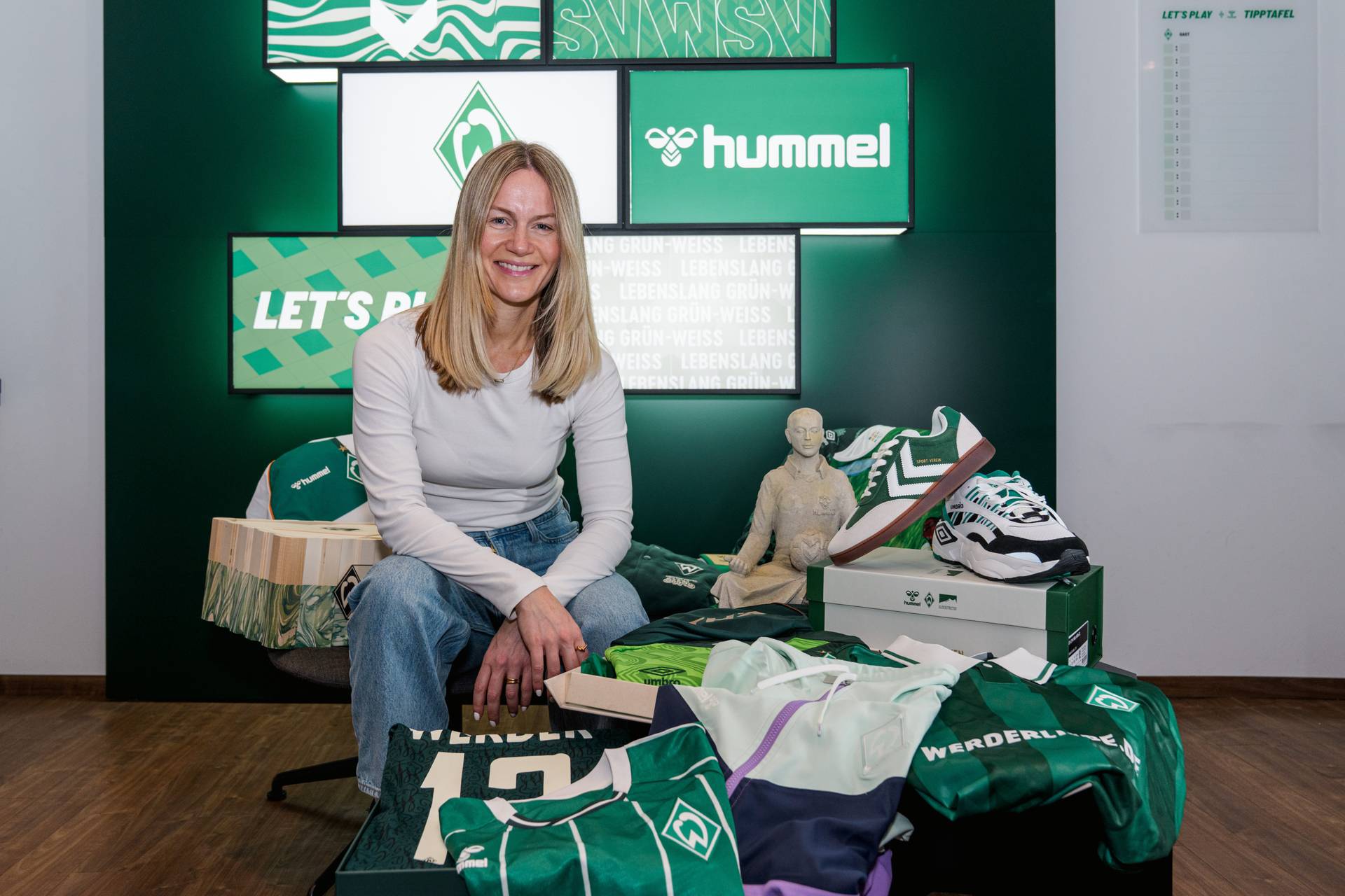 Claudia Hartung sitzt inmitten von Werder Bremen-Fanartikeln, darunter Trikots und Schuhe, mit Hummel-Logos im Hintergrund.