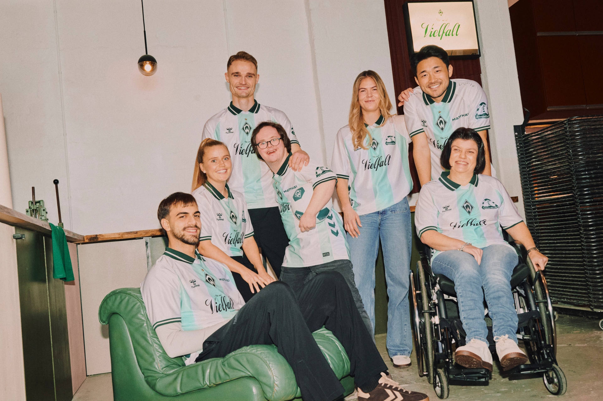 Eine Gruppe von sieben Personen in einheitlichen Uniformen, darunter eine Person im Rollstuhl, posiert gemeinsam in einer ungezwungenen Umgebung in einem Innenraum.