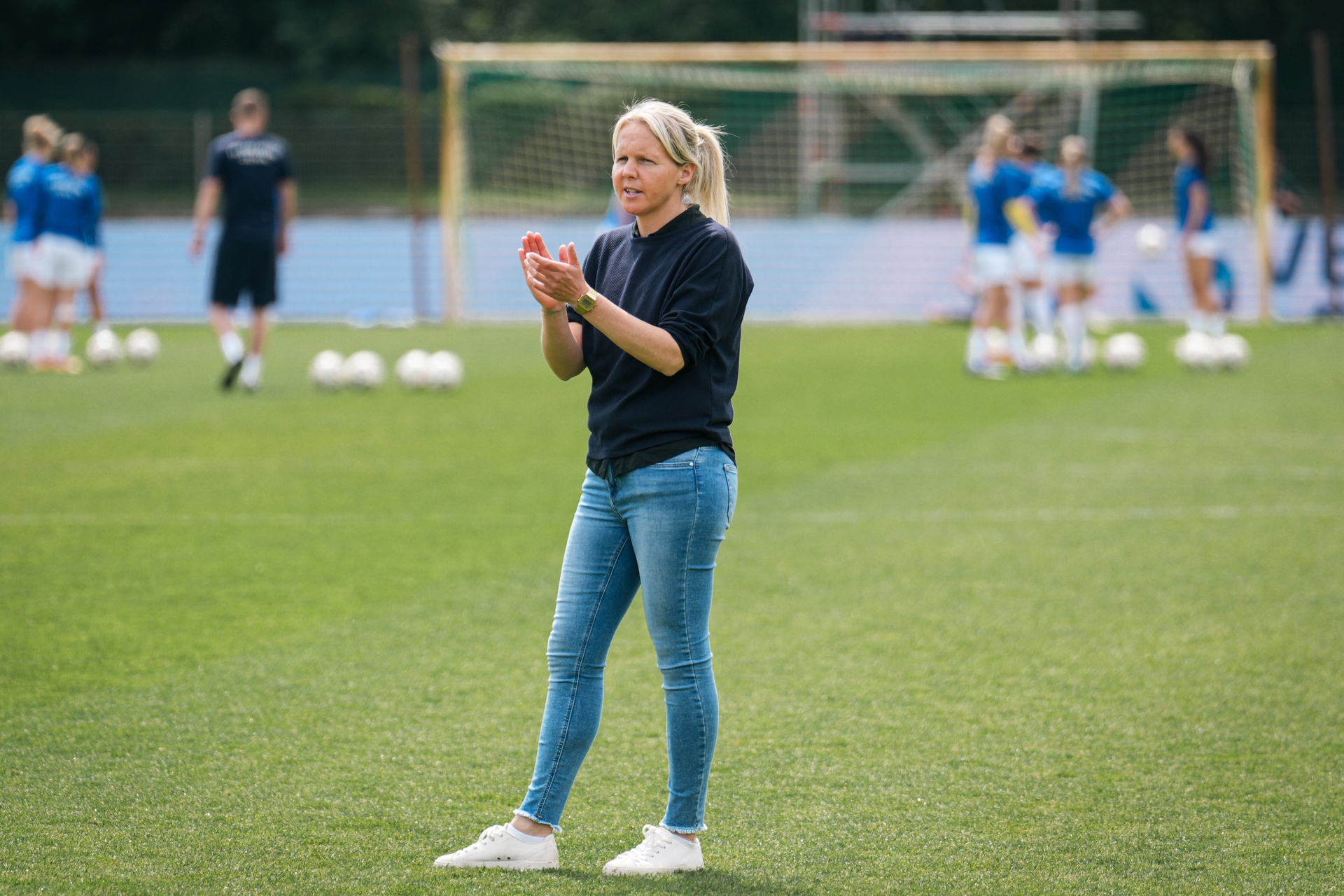 Eine Frau klatscht und scheint auf einem Fußballplatz Anweisungen zu geben. Im Hintergrund sind Spieler und Fußbälle in der Nähe eines Tores zu sehen.