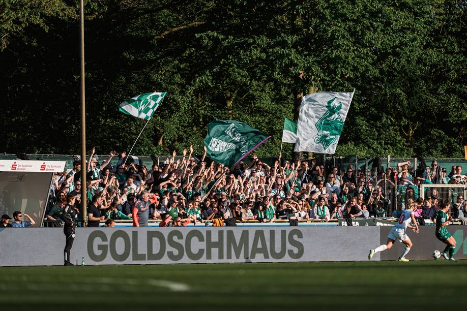 Fußballfans, die in einem vollbesetzten Stadion mit Fahnen jubeln, davor ein Schild mit der Aufschrift „GOLDSCHMAUS“. Zwei Spieler rennen über das Spielfeld.
