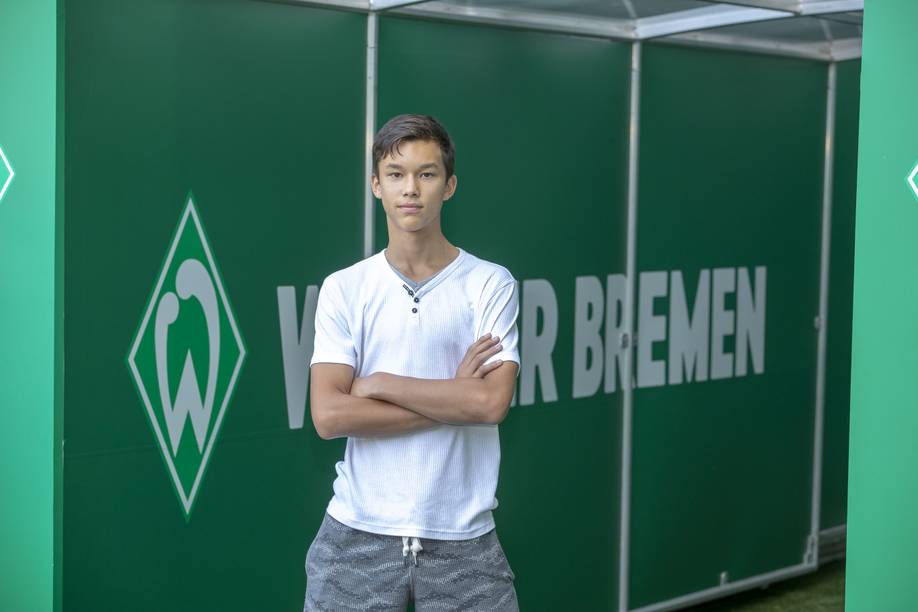 Ein junger Mann in einem weißen Hemd und gemusterten Shorts steht selbstbewusst mit verschränkten Armen vor dem grünen Hintergrund von Werder Bremen.