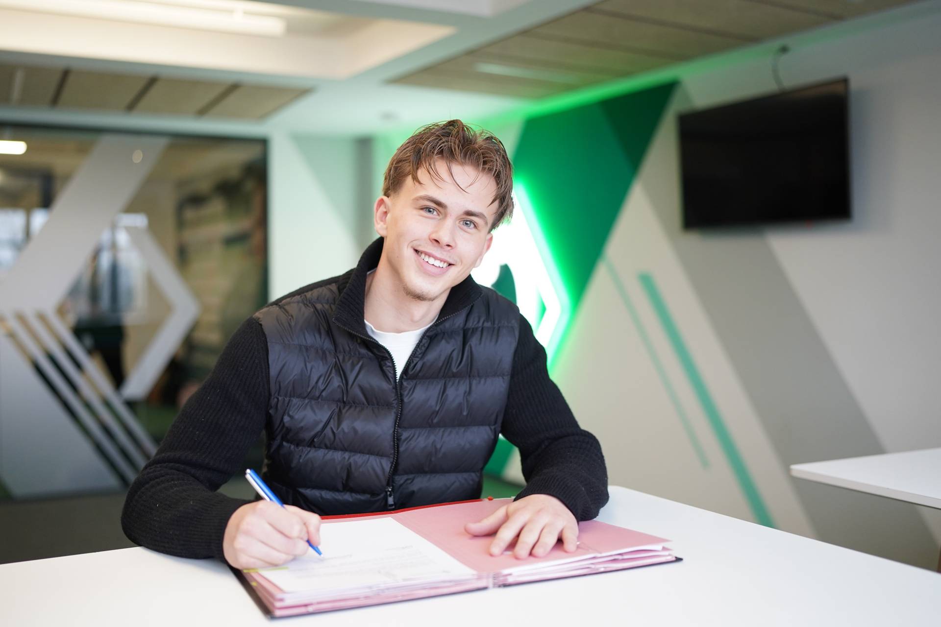 Noah Birkenpesch unterschreibt seinen Vertrag vor einer Werder-Raute.