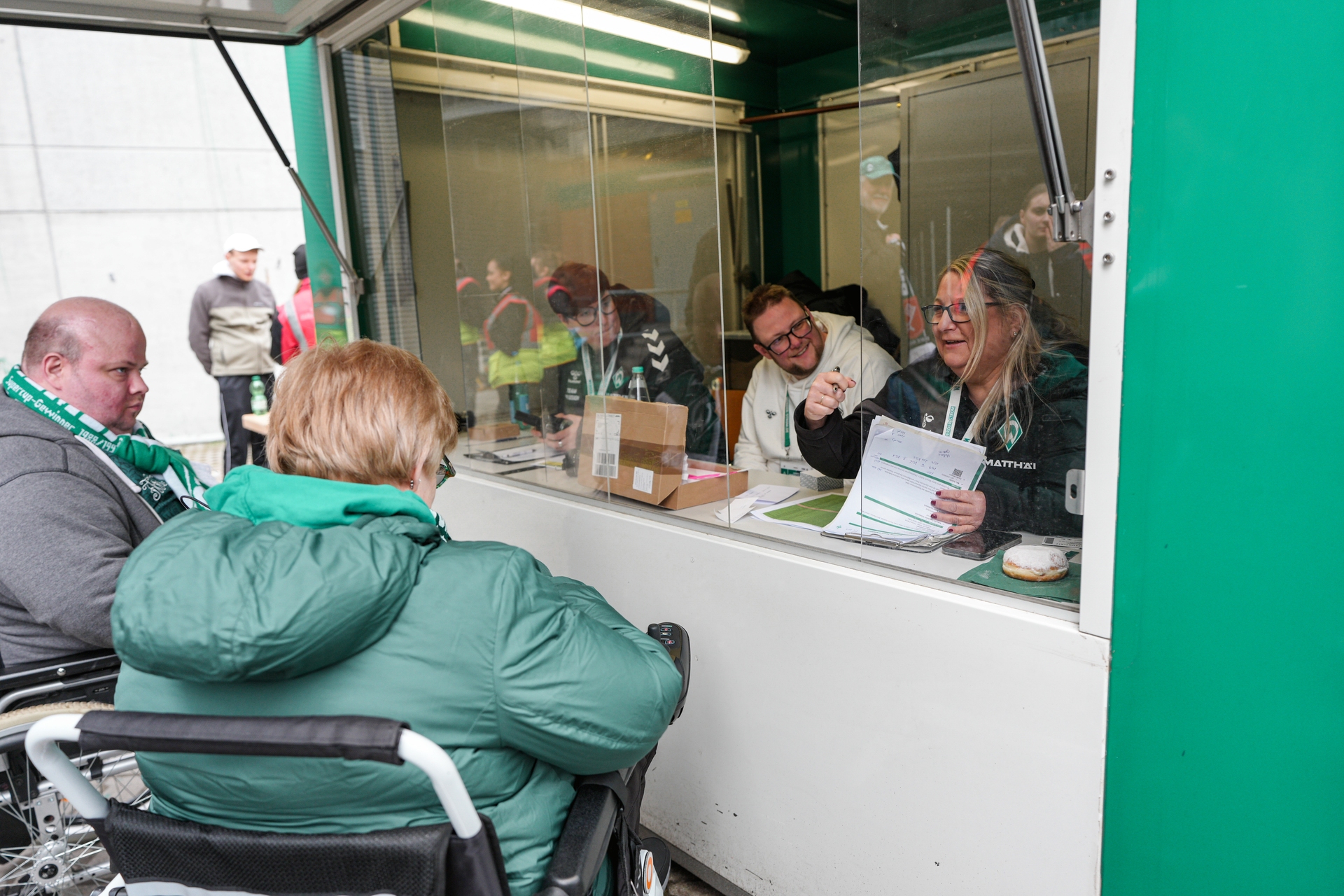 Menschen im Rollstuhl an einer Ticketbox im Austausch mit zwei Menschen hinter einer Glasscheibe sitzend.