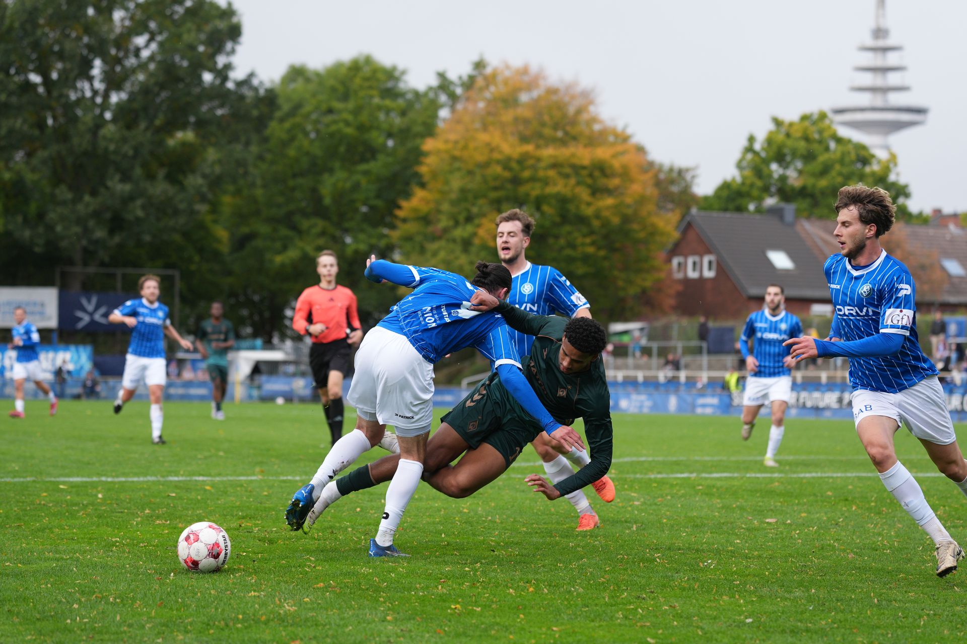 Zwei Spieler im Zweikampf. Einer fällt zu Boden, der andere hält ihn. Ein dritter Spieler läuft von rechts zum Ball, der am linken Bildrand liegt.
